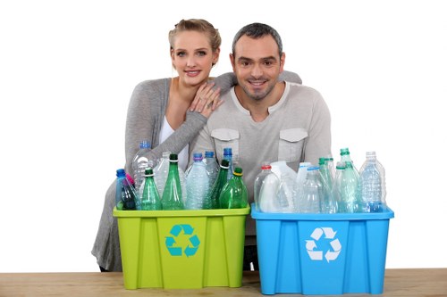 An outdoor scene featuring a man and woman standing behind a wooden table, each holding a recycling bin with a prominent recycling symbol. The woman, positioned on the left, has blonde hair tied back and is wearing a light grey cardigan, while the man on the right has short dark hair and is dressed in a light grey long-sleeved shirt. Both are smiling and appear to be sorting recyclable waste. The green recycling bin in front of the woman contains plastic bottles and containers, some with caps, made of transparent or semi-transparent plastic in various colours like green, clear, and blue. The blue recycling bin in front of the man contains additional plastic bottles and containers, also made of transparent or semi-transparent plastic. The background is plain white, highlighting the focus on the recycling bins and the individuals.