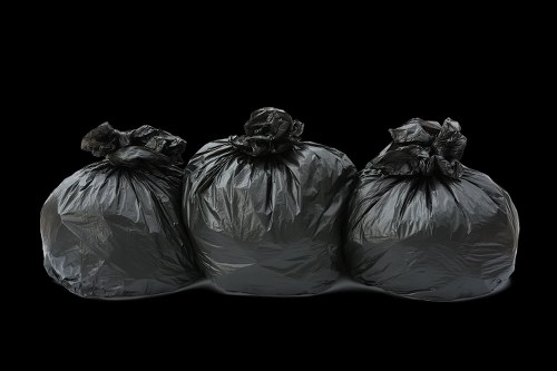Three large black rubbish bags made of thick plastic material are stacked closely together on a flat surface, with the bag openings gathered and tied at the top. The bags are slightly crumpled and reflective, showing subtle highlights due to the lighting. They are positioned in a straight line, with a dark, neutral background emphasizing their shape and texture. No other objects or environmental details are visible, focusing solely on the rubbish bags, which are typical for waste collection services in Kings Langley. The image appears professional and neutral, suitable for illustrating waste removal or rubbish collection topics on a commercial waste management website such as commercialwastekingslangley.co.uk.