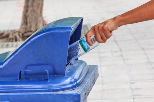 A person’s hand is seen placing a clear plastic bottle into a blue recycling bin with a hinged lid, situated on an outdoor paved surface. The bottle appears to be a standard-sized plastic container with a green cap and label, and the hand is gripping the neck of the bottle, guiding it into the bin. The recycling bin is made of durable plastic with a textured finish and has a slightly curved lid that is open to accommodate waste. In the background, there is a tree trunk visible, indicating a street or outdoor area, with the paving composed of rectangular light grey concrete tiles. The scene captures a routine act of waste disposal, reflecting environmentally responsible practices, which is relevant for local rubbish collection services in Kings Langley, near Hertfordshire. The environment is well-lit, likely during daytime, emphasizing the practical process of rubbish management managed by Commercial Waste Kings Langley.