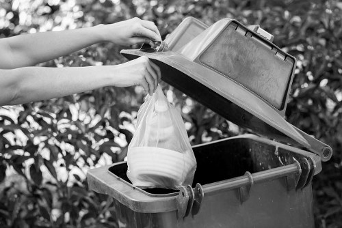 A person loading a clear plastic bag filled with white and beige waste into a black rubbish bin with a grey lid situated on a paved outdoor area, with green foliage in the background. The bin appears to be made of durable plastic, positioned on a driveway or garden path. The lid is open, revealing the bin’s interior which is shown to be empty apart from the incoming bag. The person's arms are visible, with one hand holding the plastic bag and the other steadying the bin lid. The scene reflects typical rubbish collection activities relevant to waste management services in Kings Langley, with a focus on domestic or small commercial waste disposal processes coordinated by Commercial Waste Kings Langley. Natural lighting highlights the textures of the plastic bin, the smooth surface of the bag, and the leafy backdrop, emphasizing the outdoor environment and the practical task of rubbish removal on a day-to-day basis in the local area.