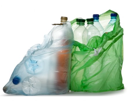 A collection of several clear plastic bottles, some with labels and caps, placed inside a green plastic refuse bag. The bottles appear to be of various sizes, with some partially crushed or flattened, and include different types of beverages. The green bag is loosely tied at the top, situated on a plain white background. The scene suggests waste collection related to beverage containers, typical of rubbish removal services provided by Commercial Waste Kings Langley. The simple composition highlights the bottles' transparency, the textures of the plastic, and the contrast between the clear bottles and the bright green bag, representing environmentally conscious waste disposal in the local area near Kings Langley postcode region.