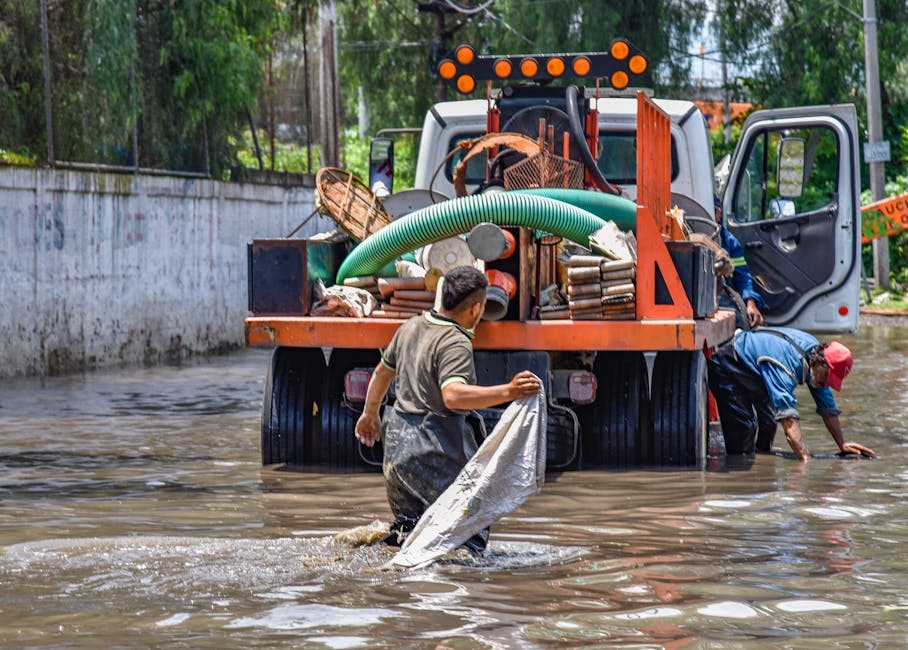 Two workers are engaged in rubbish removal or cleanup near a white utility truck parked on a flooded street. One worker, wearing a gray shirt and waterproof overalls, is holding a large white sack, wh