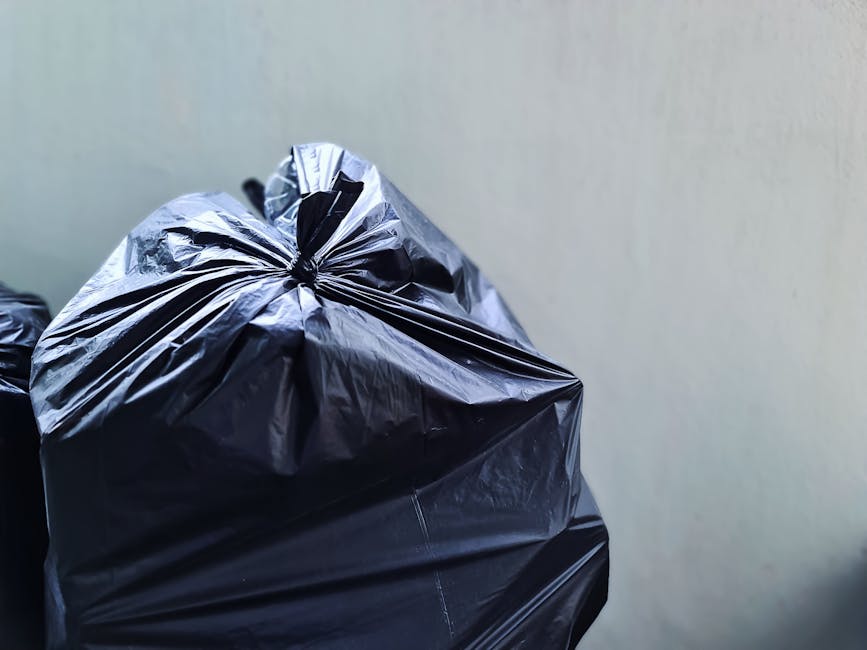 Three large black rubbish bags made of thick plastic are placed on a curbside pavement next to a black metal fence. The bags appear to be filled with waste and are leaning against each other, with the
